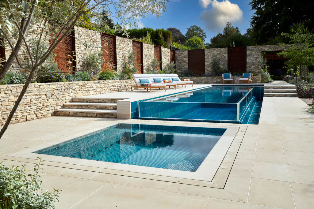 If you’re thinking of installing a swimming pool at your property, be inspired by this outdoor swimming pool design and build project in Surrey, completed earlier this year.     The property owners were looking for a bespoke swimming pool design with a boutique hotel inspired appearance, incorporating a glass infinity look and a fantastic teal glass tile colour to complement the garden design. During their pool discovery process, the property owners realised that the bespoke and challenging design required a high-end swimming pool designer and builder, which led them to Guncast Pools and Wellness. The brief was to design and build a functional outdoor swimming pool, and separate vitality pool, that could be enjoyed all year round. They wanted a modern shape and design that fitted well in the garden space and made use of the garden’s natural levels.  The infinity edge pool design serves as both an elegant water feature, visible from the house at all times, and a stylish swimming pool used by the owners for relaxing, exercising and socialising / entertaining. The outdoor rectangular-shaped, infinity edge pool measures 12m x 3m, with a consistent depth of 1.2m and is maintained at a comfortable 29°C. The deck-level square vitality pool measures 3.5m x 3.5m, with a 0.98m depth. It is kept at a soothing 34-35°C and comfortably fits 6-8 people.    The property owner said about working with Guncast Pools and Wellness: “Through the design and specification process, it was clear that the Guncast team were experienced and very detailed in terms of the pool requirements.  The proposal and scope were very thorough and gave us confidence that the project was achievable.  “Overall, despite some challenging elements, the team have been very competent and the finished project is outstanding both in terms of appearance but also function and practicality.”  If you’re thinking of a swimming pool for your property, contact the Guncast design team to get started.  Be inspired – take a look at our portfolio.
