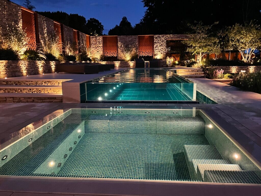 vIf you’re thinking of installing a swimming pool at your property, be inspired by this outdoor swimming pool design and build project in Surrey, completed earlier this year.     The property owners were looking for a bespoke swimming pool design with a boutique hotel inspired appearance, incorporating a glass infinity look and a fantastic teal glass tile colour to complement the garden design. During their pool discovery process, the property owners realised that the bespoke and challenging design required a high-end swimming pool designer and builder, which led them to Guncast Pools and Wellness. The brief was to design and build a functional outdoor swimming pool, and separate vitality pool, that could be enjoyed all year round. They wanted a modern shape and design that fitted well in the garden space and made use of the garden’s natural levels.  The infinity edge pool design serves as both an elegant water feature, visible from the house at all times, and a stylish swimming pool used by the owners for relaxing, exercising and socialising / entertaining. The outdoor rectangular-shaped, infinity edge pool measures 12m x 3m, with a consistent depth of 1.2m and is maintained at a comfortable 29°C. The deck-level square vitality pool measures 3.5m x 3.5m, with a 0.98m depth. It is kept at a soothing 34-35°C and comfortably fits 6-8 people.    The property owner said about working with Guncast Pools and Wellness: “Through the design and specification process, it was clear that the Guncast team were experienced and very detailed in terms of the pool requirements.  The proposal and scope were very thorough and gave us confidence that the project was achievable.  “Overall, despite some challenging elements, the team have been very competent and the finished project is outstanding both in terms of appearance but also function and practicality.”  If you’re thinking of a swimming pool for your property, contact the Guncast design team to get started.  Be inspired – take a look at our portfolio.