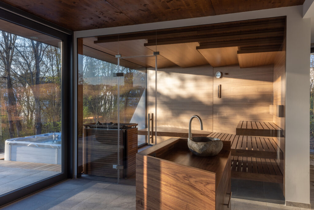 Take a look at this stunning wellness space created by KLAFS in Germany for a high-end residential property, in Arnstein, Germany. Featuring an impressive steam bath, a soothing dark wood sauna, and a meditative anteroom, this wellness space offers the perfect environment to regenerate both body and mind. The client was looking for multi-functional, bespoke design, and quickly decided that KLAFS should be involved with the project planning and installation, together with the project architects and builders. Experts at the KLAFS showroom in Schwäbische Hall, gave comprehensive guidance showed the client and architects the extensive range of KLAFS wellness products and accessories. A sauna with a view Following the consultation with the KLAFS team, the clients settled on a high-quality, individually designed KLAFS Sauna PROFI. The customised cabin was installed in a solid walnut finish. The KLAFS Sauna features a large glass front, giving a panoramic view of the client’s stunning garden space. With plenty of natural light during the day and warm indirect LED lighting at night, this wellness space was built to offer a soothing atmosphere around the clock. Whilst the dark American walnut of the sauna lends a cosy, comfortable atmosphere, the generous panorama window provides transparency and lightness. This contrasting interplay makes the sauna a spacious, light, and airy oasis of relaxation, which loses none of its feeling of security thanks to the warmth and natural beauty of the wooden walls and benches. The cabin’s benches, with their concealed mountings, give the appearance of floating and are a design highlight of this bespoke wellness project, complimented by the stepped ceiling. Opulent steam bath design A testimony to the meticulous planning and craftsmanship of this project can be found in the steam bath which adjoins the sauna, and was built into a beautiful, domed room. The entire steam room is fitted with luxury golden-brown Bisazza mosaic tiles and features a gold recess. The curves of the seating area and the recess posed a particular challenge for the builders - and the effort was more than worth it. The stylish and harmonious design helps to give every user of this steam bath a unique and soothing experience. Atmospheric sauna lighting and a 'starlit' steam room In addition to natural daylight, the sauna features indirect LED lighting on the backrest, which complements the perfectly coordinated lighting concept and creates the right atmosphere for every mood. The interior of the sauna and the relaxation area are designed with matching material, creating a harmonious effect between the two wellness spaces. The relaxation area (anteroom) is equipped with gentle, atmospheric light sources and offers the ideal atmosphere to simply unwind after a relaxing sauna bath. The unique steam bath space is complemented by the sparkling filigree stars of the POLAR STARRY HEAVEN design installed on the ceiling, combined with gentle, indirect LED lighting. Guncast offers several lighting options for your wellness area and can even account for spaces with low levels of natural light with solutions such as Sun Meadow UV Lighting. Find out more about by contacting our design team. Talk to Guncast about professional and home wellness projects The KLAFS team worked with a team of architects and builders to create this relaxation oasis. Guncast also works closely with architects, property developers, and project managers, to create stunning swimming pools and wellness projects for domestic or commercial properties. We supply KLAFS turnkey and bespoke designed saunas, steam rooms, and wellness products to help you create your own exclusive oasis, designed, and built to suit your clients’ space. Contact the Guncast team or make an appointment to visit the Manchester showroom.