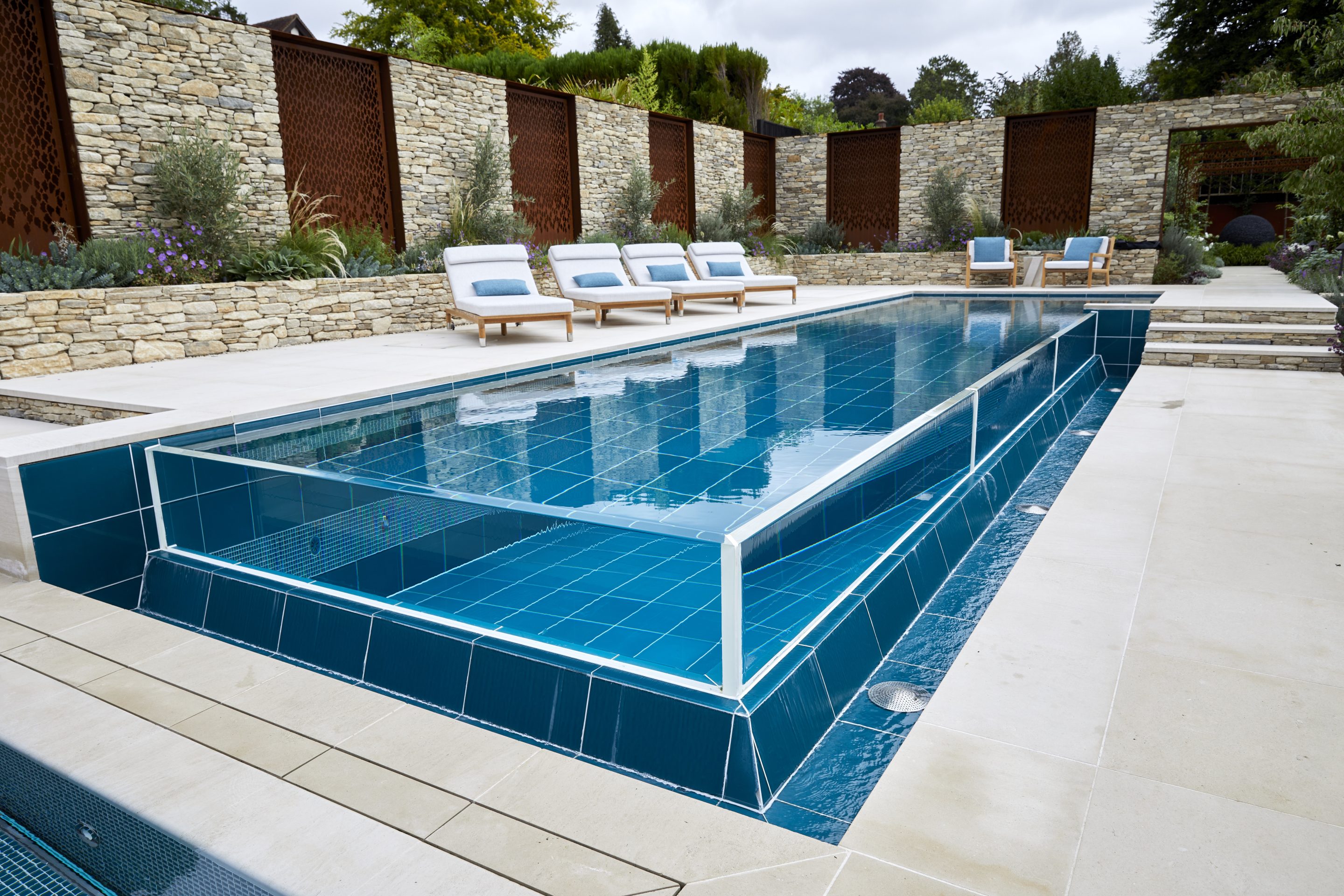 A distinctive outdoor infinity pool design from a project in Surrey. Pool is crystal clear blue, face on at an angle leaning to the left with deck chairs to the left and a path with steps to the right.